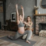 Pregnant woman doing yoga poses with a trainer in cozy indoor setting.