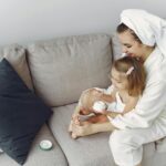 A mother and daughter in bathrobes bonding on a couch, applying moisturizer together.