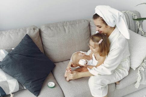 A mother and daughter in bathrobes bonding on a couch, applying moisturizer together.