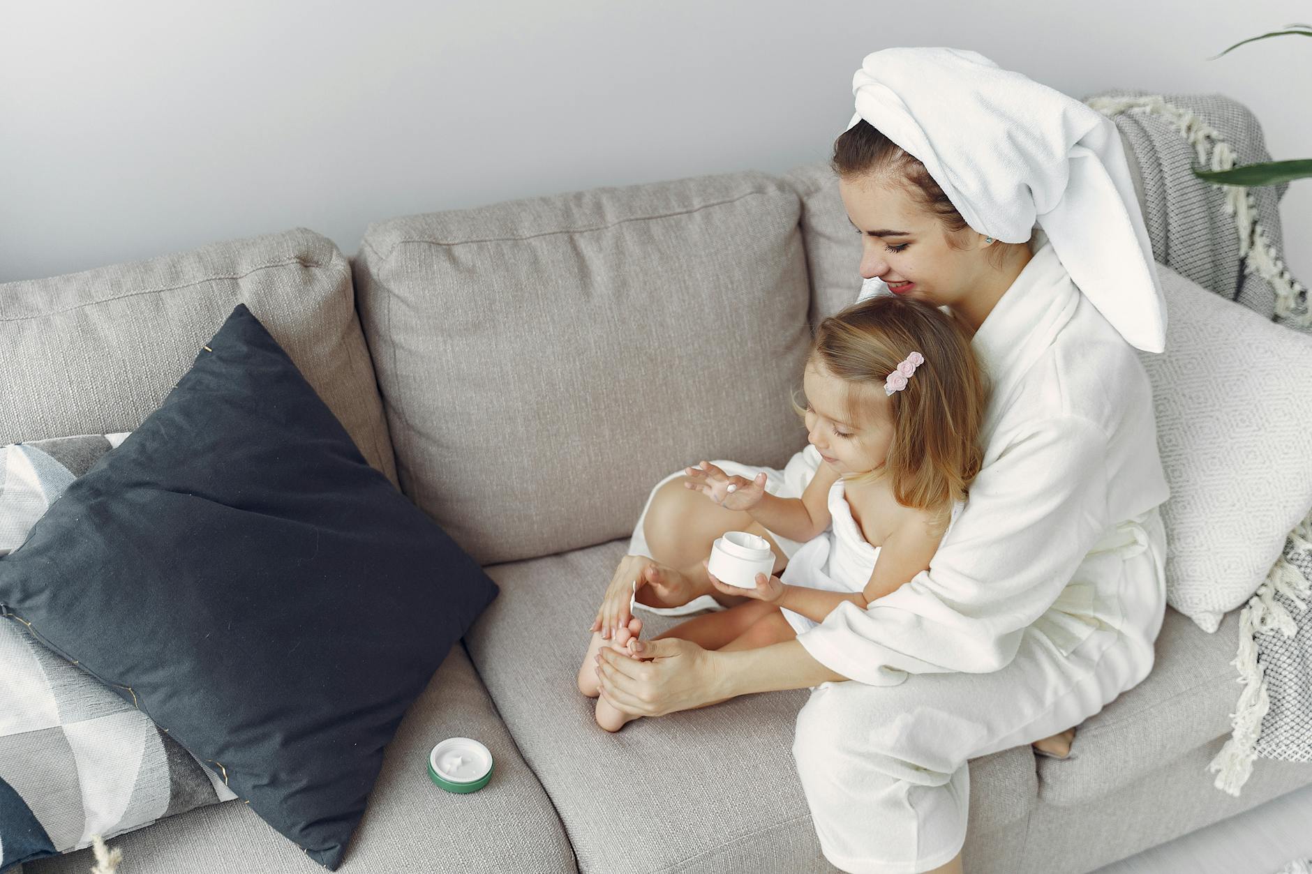 A mother and daughter in bathrobes bonding on a couch, applying moisturizer together.