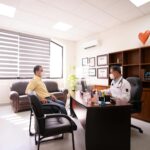 A patient consults with a masked doctor in a well-lit, modern office. Safety measures in place.