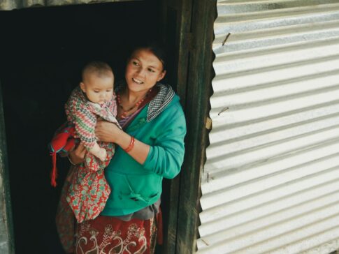 Smiling mother holds her baby while standing at the doorway of a rustic home, showcasing familial bond.