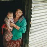 Smiling mother holds her baby while standing at the doorway of a rustic home, showcasing familial bond.