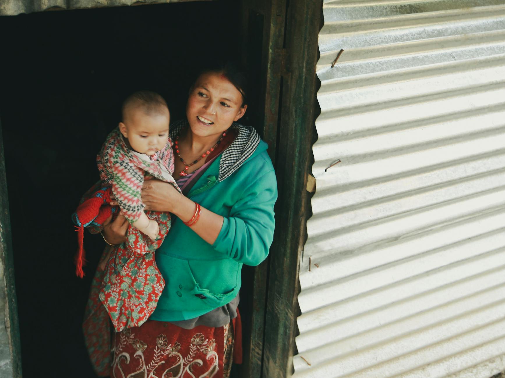 Smiling mother holds her baby while standing at the doorway of a rustic home, showcasing familial bond.
