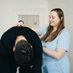 A physiotherapist smiling while assisting a patient with a back exercise in a clinic.