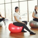 Positive pregnant ladies sitting on fit balls during prenatal training in spacious light gym at daytime and smiling while looking at camera