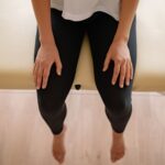 A woman sitting barefoot on a massage table indoors, captured from above.