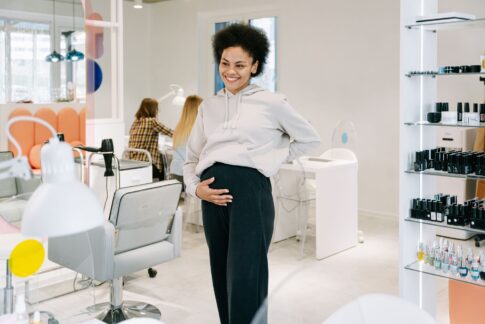A smiling pregnant woman stands in a stylish nail salon, showcasing joy and relaxation.
