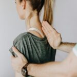 Back view of crop unrecognizable osteopath in uniform and wristwatch checking up back of slim female patient in casual wear on white background