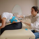 A physiotherapist applies kinesiology tape to a young woman's back during a therapy session.