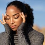 Close-up of a woman in a gray sweater holding her temples, experiencing a headache outdoors in the sunlight.