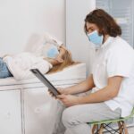 Doctor consulting patient wearing face masks in a medical examination room, discussing healthcare plan.