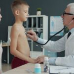 A doctor uses a stethoscope to examine a young boy with his mother present in a medical office.
