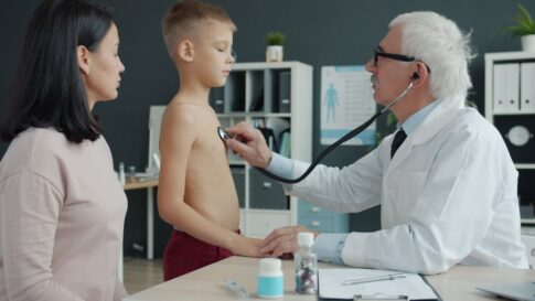 A doctor uses a stethoscope to examine a young boy with his mother present in a medical office.