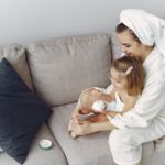 A mother and daughter in bathrobes bonding on a couch, applying moisturizer together.