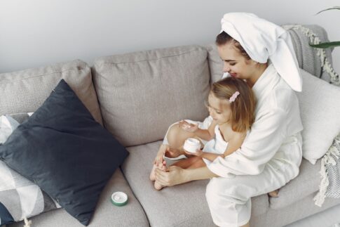 A mother and daughter in bathrobes bonding on a couch, applying moisturizer together.