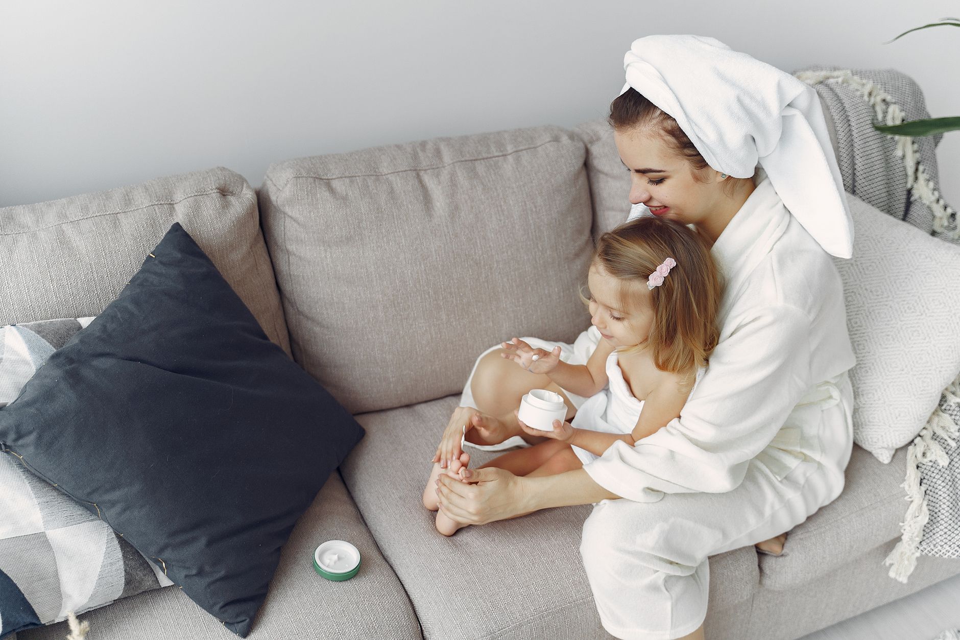 A mother and daughter in bathrobes bonding on a couch, applying moisturizer together.