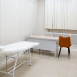 A minimalist medical examination room featuring a desk, chairs, and an examination bed in a sterile, neutral setting.