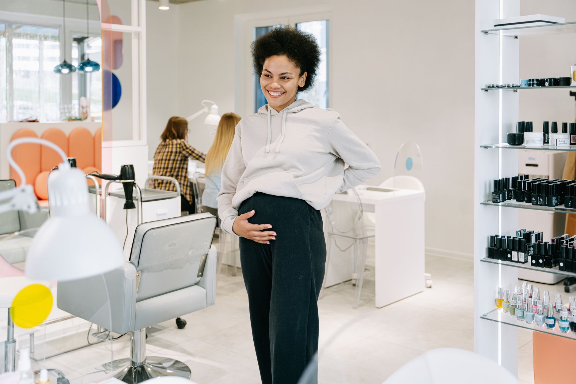 A smiling pregnant woman stands in a stylish nail salon, showcasing joy and relaxation.